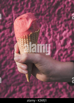 La mano di un ragazzo in possesso di un gelato alla fragola cono su uno sfondo rosa Foto Stock