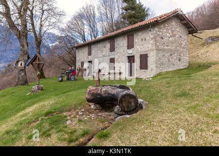 Paio di escursionisti sedersi sulle panchine al di fuori del Nusieda alta capanna, in primo piano la grande fontana scolpita in un tronco di legno. Monti del Sole, Parco Nazionale Dolomiti Bellunesi, Belluno, Veneto, Italia Foto Stock