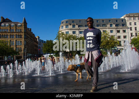 Manchester, Regno Unito - 15 August, 2017: l uomo e il suo cane a piedi mediante la Piccadilly Gardens fontane ad acqua nel centro della città di Manchester in una giornata di sole Foto Stock