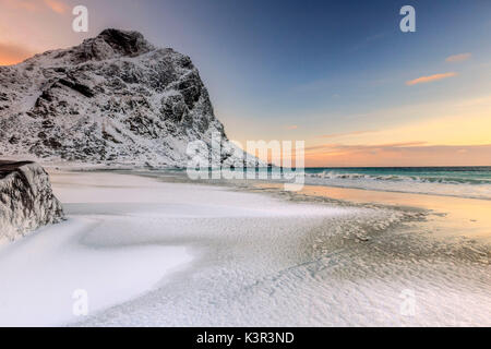 Onde avanza verso la riva del mare circondato da vette innevate all'alba. Uttakleiv Isole Lofoten in Norvegia Europa Foto Stock