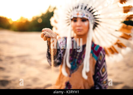 Bella donna in un abito di indiani americani gioca con la sabbia sul tramonto. Giovane ragazza nel copricapo fatto di piume di uccelli selvatici Foto Stock