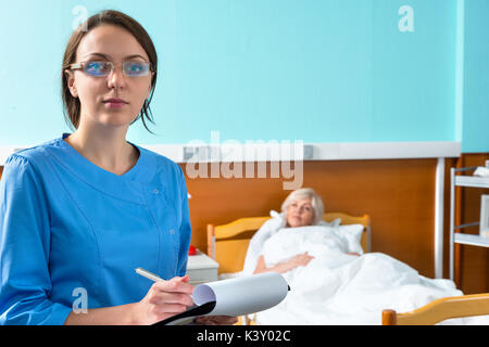 Giovani femmine infermiere o medico in uniforme di indossare una maschera e occhiali facendo alcune note in relazione al clipboard mentre visitano il suo paziente malato in corsia Foto Stock