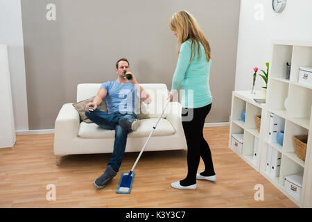 Donna tergi pavimento mentre l'uomo sul divano di bere alcolici Foto Stock