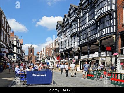 Edifici e negozi lungo Bridge Street con la chiesa di St Peters verso la parte posteriore e un cafe' sul marciapiede in primo piano, Chester, Cheshire, Inghilterra, Regno Unito, Weste Foto Stock