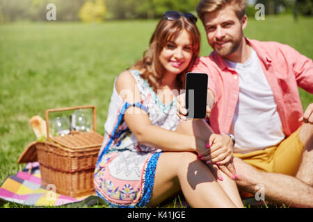 Una foto di una giovane coppia felice picnic sul prato. L'uomo sta tenendo il suo nuovissimo smartphone. Foto Stock