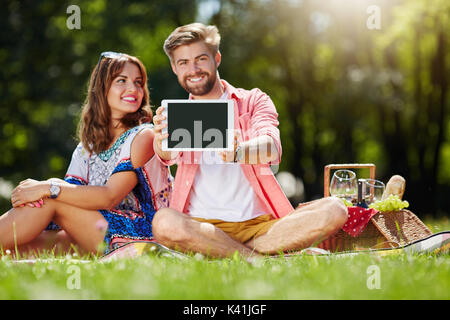 Una foto di una giovane coppia felice picnic sul prato. l uomo sta tenendo il suo nuovissimo tablet. Foto Stock