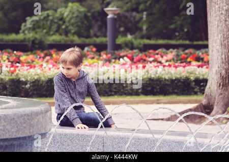 Bambino seduto vicino alla fontana nel parco della città Foto Stock