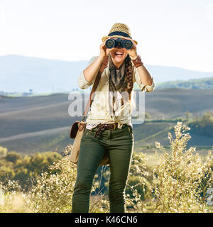 Alla scoperta di magiche vedute della Toscana. adventure donna escursionista in hat godendo la Toscana vista guardando nella telecamera attraverso il binocolo Foto Stock