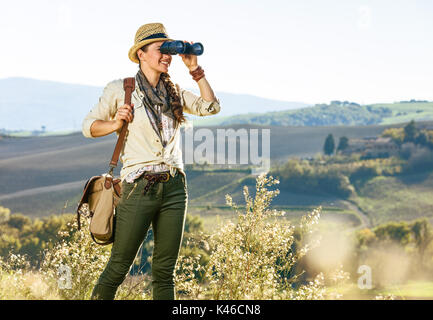 Alla scoperta di magiche vedute della Toscana. donna sana escursionista in hat con sacchetto sulla Toscana Escursione esaminando la distanza attraverso il binocolo Foto Stock