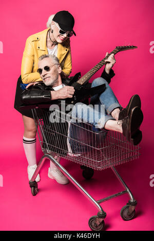 Sorridente bella donna senior appoggiata al carrello della spesa con uomo senior di suonare una chitarra elettrica Foto Stock