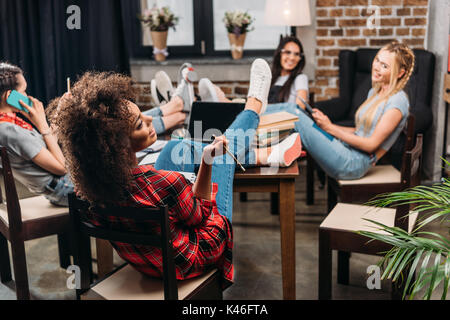 Gruppo multietnico di studenti seduti su sedie, studiando e utilizzando dispositivi digitali Foto Stock