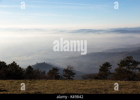 Una vista della città di Assisi e san.francesco chiesa sopra un mare di nebbia, con lontane colline e montagne sullo sfondo, e alcuni alberi in primo piano Foto Stock