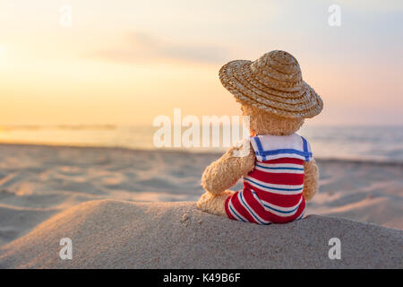 Vista posteriore del piccolo orsacchiotto, usura nostalgico del costume da bagno a righe e cappello di paglia, seduto presso la spiaggia di sabbia e godere di sunny atmosfera serale in mare (copia spa Foto Stock