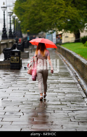 Vista posteriore femminile che cammina con ombrello luminoso sotto la pioggia a Londra. Marciapiede bagnato Foto Stock