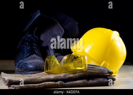 Indumenti da lavoro per un lavoratore edile. Casco, guanti, scarpe e occhiali da sole su di un tavolo di legno. Sfondo nero. Foto Stock