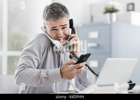 Sottolineato imprenditore disperato lavorando nel suo ufficio e avente le chiamate multiple, egli è in possesso di due telefoni e di un telefono cellulare, la gestione del business con Foto Stock