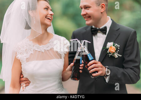 La vista ravvicinata della sorridenti sposi novelli clincking le bevande nel parco. Foto Stock