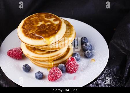 Pila di pancake con lamponi e mirtilli rivestite con zucchero a velo sulla piastra bianca e sfondo nero Foto Stock