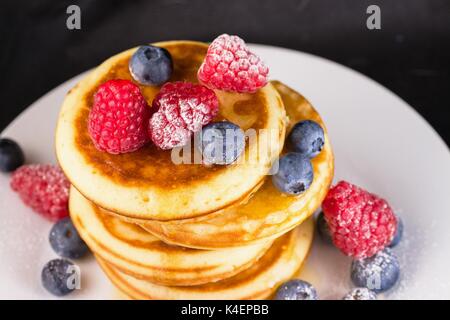 Lo zucchero in polvere rivestita di lamponi e mirtilli in sciroppo sulla parte superiore della pila di pancake Foto Stock