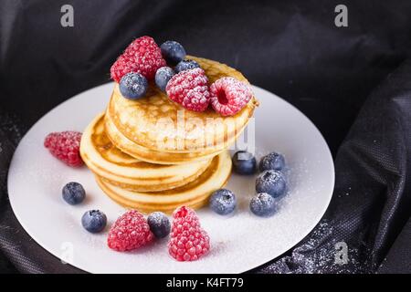 Pila di pancake con lamponi e mirtilli rivestite con zucchero a velo sulla piastra bianca e sfondo nero Foto Stock