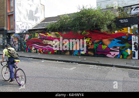 Sfocata ciclista la sua faccia nascosta passato in bicicletta a vivaci e colorati luminosamente street murale su sclater street vicino a Brick Lane, London, Regno Unito Foto Stock