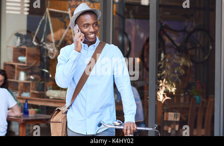 Elegantemente vestito giovane africano a parlare sul suo cellulare mentre si cammina con la sua bicicletta giù per una strada di città Foto Stock