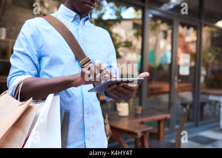 Primo piano di un elegante giovane africano utilizzando una tavoletta digitale mentre in piedi con le borse della spesa su una strada di città Foto Stock