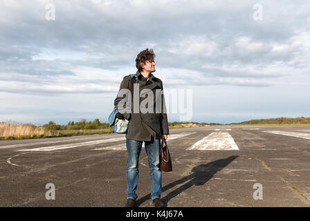 Giovane uomo in blue jeans e cappotto grigio permanente sulla pista di andare in vacanza in aereo con pelle marrone borsa in mano. aeromobili bianco sullo sfondo Foto Stock