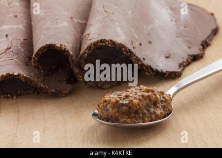Tre fette di cuocere la carne di manzo britannica con una crosta pepato accanto a un cucchiaino colmo di senape integrali su una tavola di legno. Foto Stock