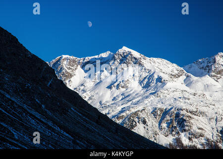La luna appare sopra le cime innevate nel cielo blu Julierpass Albula Distretto Cantone dei Grigioni Svizzera Europa Foto Stock