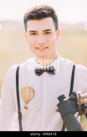 Close-up ritratto verticale del sorridente lo sposo in camicia bianca decorata wth aria-palloncino e il filtro bow tie tenendo il binocolo. Foto Stock