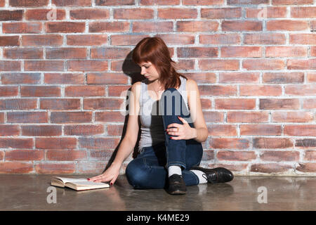 Giovane e bella capelli rossi donna in jeans con libro seduti sul pavimento vicino a un muro di mattoni. attentamente la lettura di un libro interessante. Foto Stock