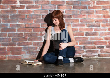 Giovane e bella capelli rossi donna in jeans con libro seduti sul pavimento vicino a un muro di mattoni. attentamente la lettura di un libro interessante. Foto Stock