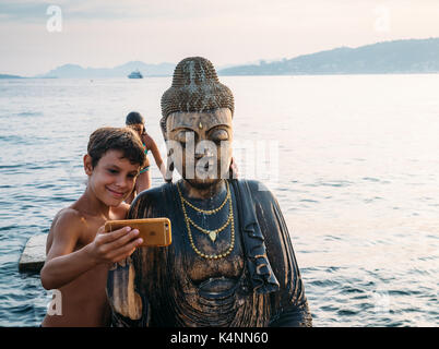 Un giovane ragazzo prende un selfie con una statua di Budda su una spiaggia in Juan les Pins, Francia Foto Stock