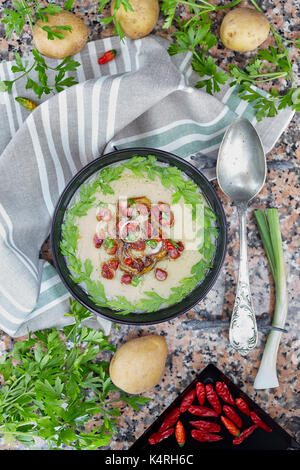 Cremosa zuppa di patate in nero ciotola decorata con pezzi di pancetta arrosto, cipolline, giallo pepe, peperoncino e prezzemolo, vista top shot. Foto Stock