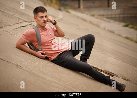 Il ragazzo ascolta musica e giace su lastre in calcestruzzo. Il resto sulla strada. ha un piccolo zaino con lui. moda giovane. Foto Stock