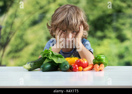 Carino piccolo ragazzo seduto al tavolo, infelice con il suo pasto vegetale, cattive abitudini alimentari, nutrizione e alimentazione sana nozione Foto Stock