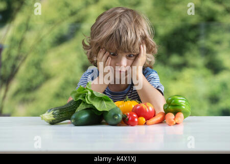 Carino piccolo ragazzo seduto al tavolo, infelice con il suo pasto vegetale, cattive abitudini alimentari, nutrizione e alimentazione sana nozione Foto Stock