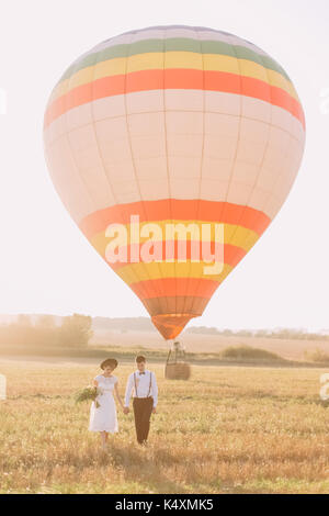 La verticale a tutta lunghezza Ritratto di felice sposi novelli azienda habds e camminare durante il tramonto nel campo in background della enorme colorati airballoon. Foto Stock