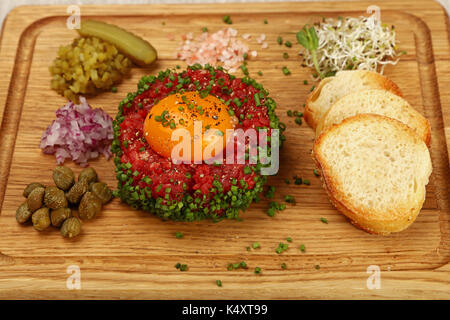 Una porzione di materie le carni bovine macinate tartare di carne di manzo con tuorlo d'uovo, baguette di pane, verde erba cipollina, cipolle e capperi su tavola di legno, vicino, angolo alto Foto Stock