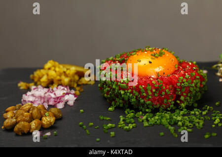 Una porzione di materie le carni bovine macinate tartare di carne di manzo con tuorlo d'uovo, verde erba cipollina, cipolle, cetrioli e capperi su nero ardesia bordo, vicino, angolo alto Foto Stock