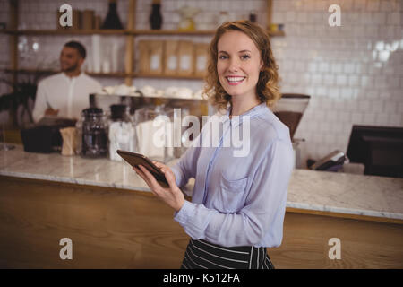 Sorridente giovane cameriera con tavoletta digitale mentre in piedi dal contatore al coffee shop Foto Stock