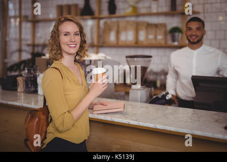 Ritratto di giovane sorridente femmina azienda cliente monouso tazza di caffè al bancone contro il cameriere in caffetteria Foto Stock