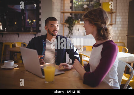 Giovani amici di parlare durante la seduta con il computer portatile e drink presso la caffetteria Foto Stock