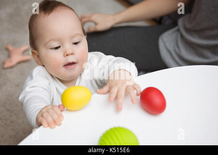 Nove mesi Baby girl cercando di agguantare il giocattolo colorato uova sul tavolo bianco; madre seduta al suo fianco Foto Stock