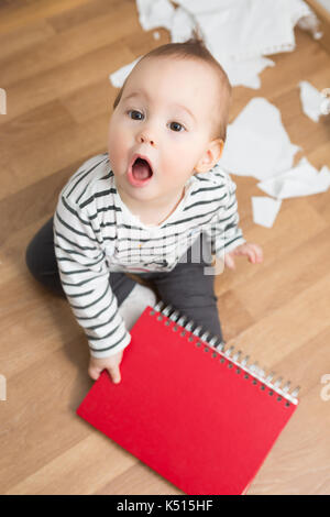 Dieci mesi Baby girl giocando con documenti e la cartella della carta sul pavimento; lo strappo della carta e la creazione di un pasticcio in camera Foto Stock