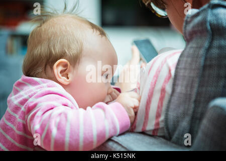 Dieci mesi Baby girl cercando di ottenere di sua madre attenzione; madre è seduta su un divano texting messaggi sullo smartphone Foto Stock