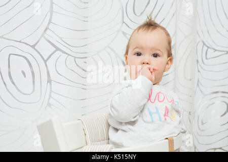 Undici mesi baby ragazza seduta in uno sgabello facendo facce buffe nella fotocamera e mettendo il dito in bocca Foto Stock