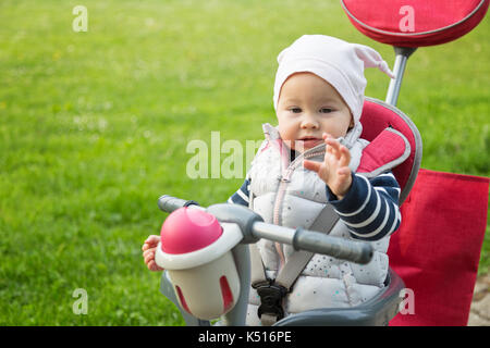 Un anno di età baby ragazza seduta in un rosso e grigio triciclo nel parco, contro il soleggiato di erba; con la sua mano alzata e agitando Foto Stock