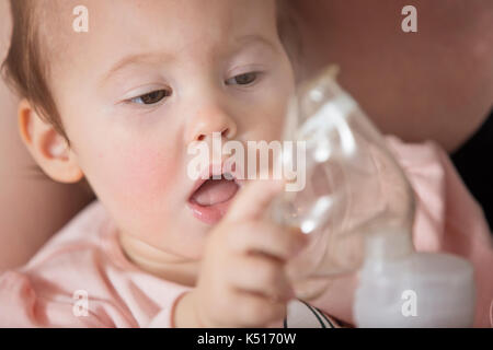 Un anno di età Baby girl tenendo l'inalatore, sua madre mentre tiene il suo nei bracci e consolante il bambino malato Foto Stock